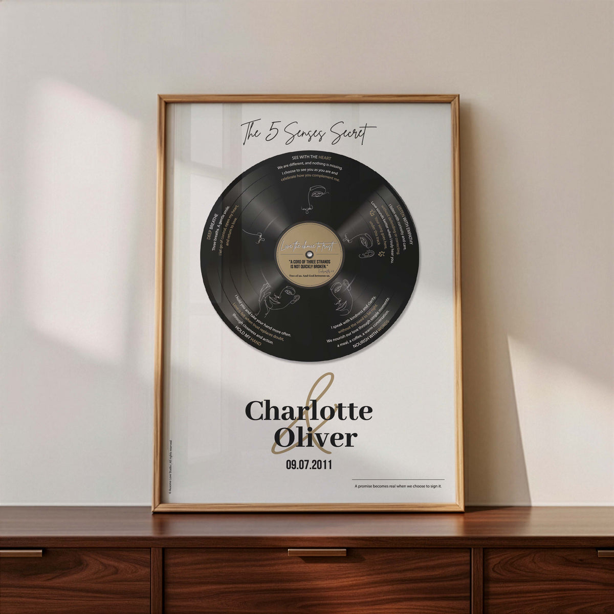 Personalised couple print with a black vinyl record on a cream background, featuring five sense-themed love messages and line-art faces, the names Charlotte & Oliver and the date 09.07.2011 below, in a timber frame resting on a wooden sideboard.