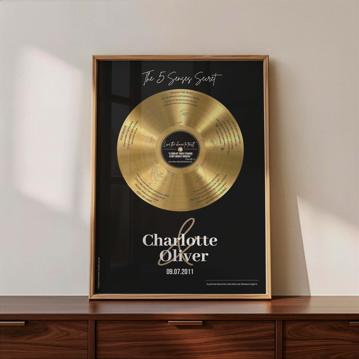 Personalised couple print with a gold vinyl record on a black background, featuring five sense-themed love messages and line-art faces, the names Charlotte & Oliver and the date 09.07.2011 below, in a timber frame resting on a wooden sideboard.