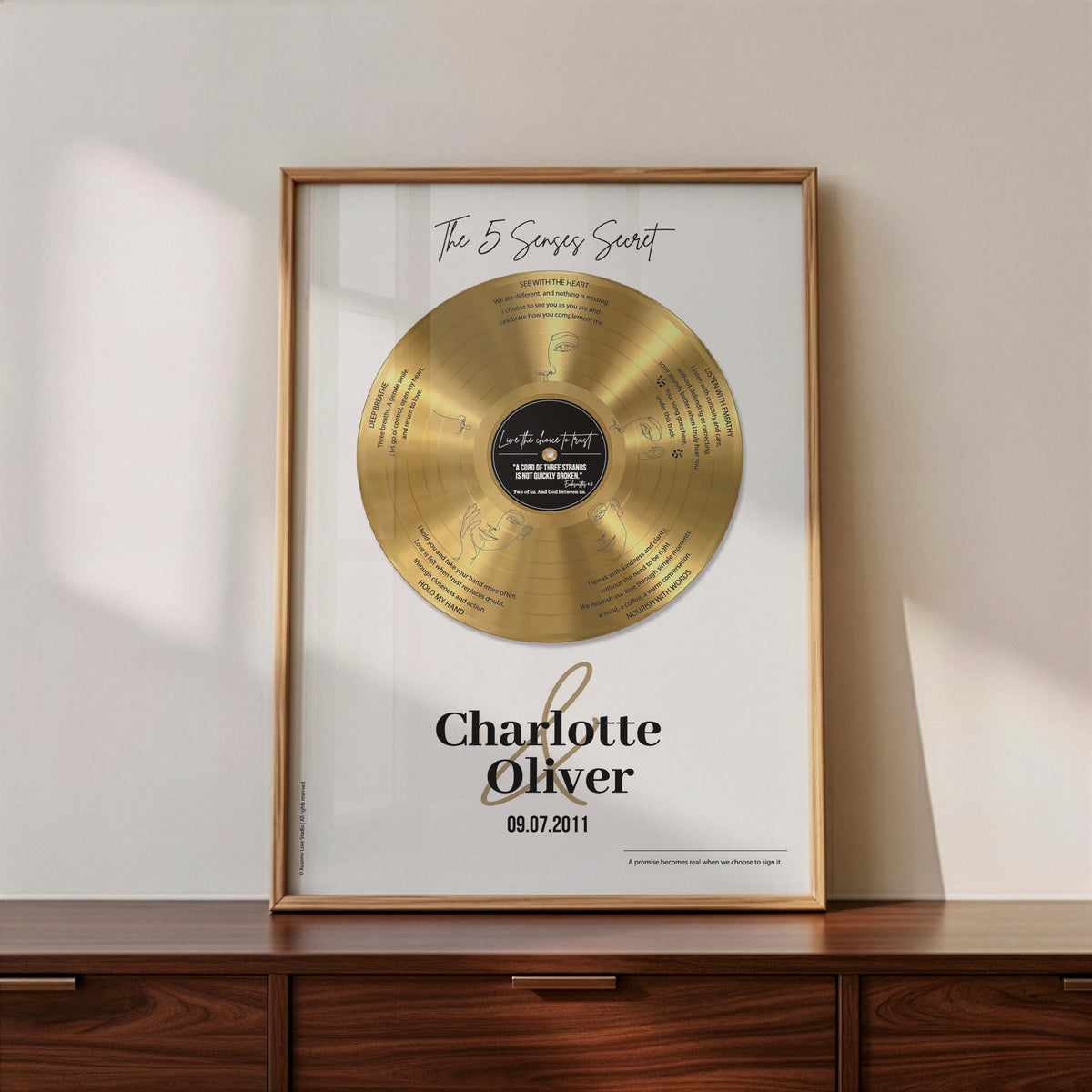 Personalised couple print with a gold vinyl record on a cream background, featuring five sense-themed love messages and line-art faces, the names Charlotte & Oliver and the date 09.07.2011 below, in a timber frame resting on a wooden sideboard.