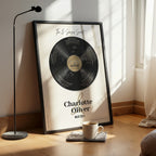 Personalised couple print with a black vinyl record on a cream background, featuring five sense-themed love messages and line-art faces, the names Charlotte & Oliver and the date 09.07.2011 below, in a black frame leaning against a sunlit wall beside a coffee cup.
