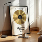 Personalised couple print with a gold vinyl record on a cream background, featuring five sense-themed love messages and line-art faces, the names Charlotte & Oliver and the date 09.07.2011 below, in a black frame leaning against a sunlit wall beside a coffee cup.