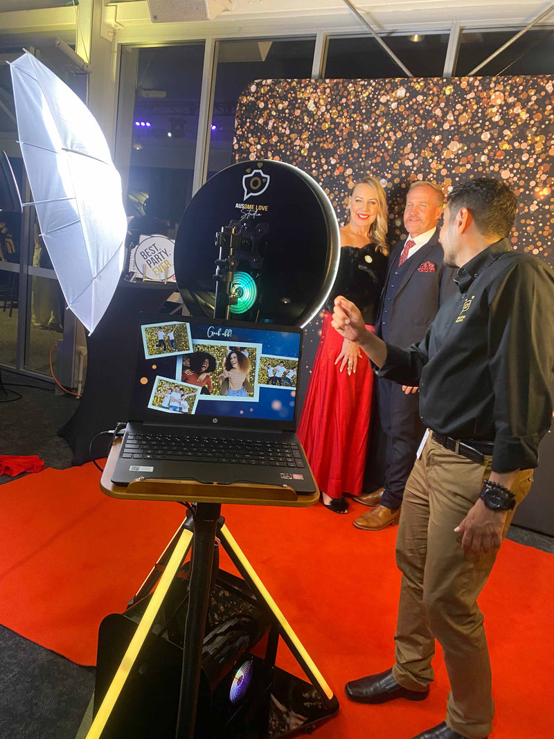 The Ausome Love Studio attendant helping guests at a photo booth during a formal event, with professional umbrella lighting, a gold sequin backdrop and red carpet