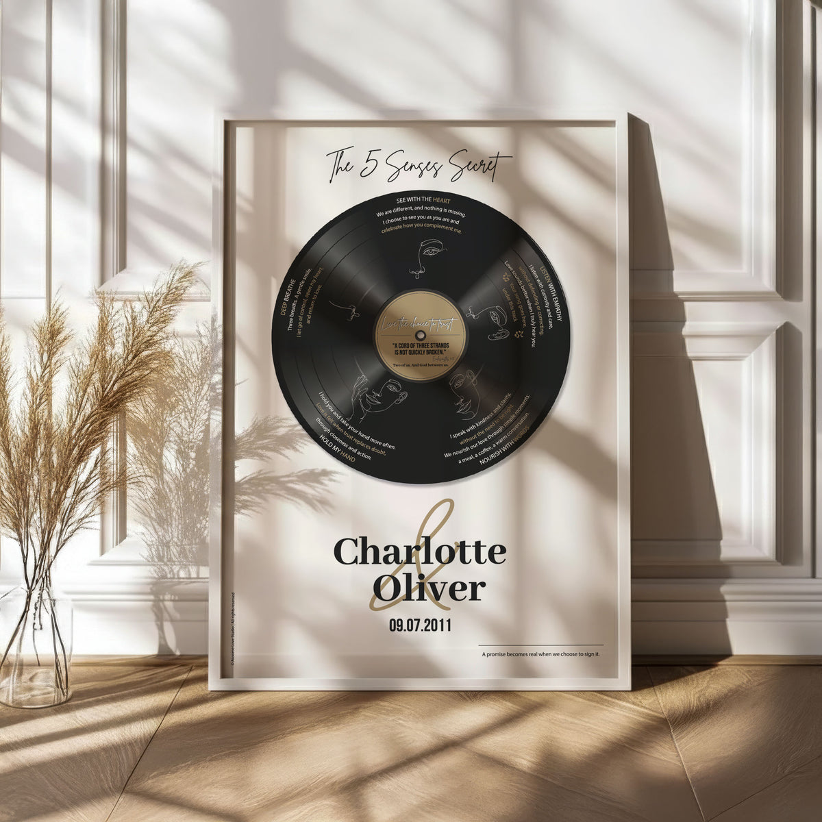 Personalised couple print with a black vinyl record showing five sense-themed love messages and line-art faces, the names Charlotte & Oliver and the date 09.07.2011 below, framed in white. Main image